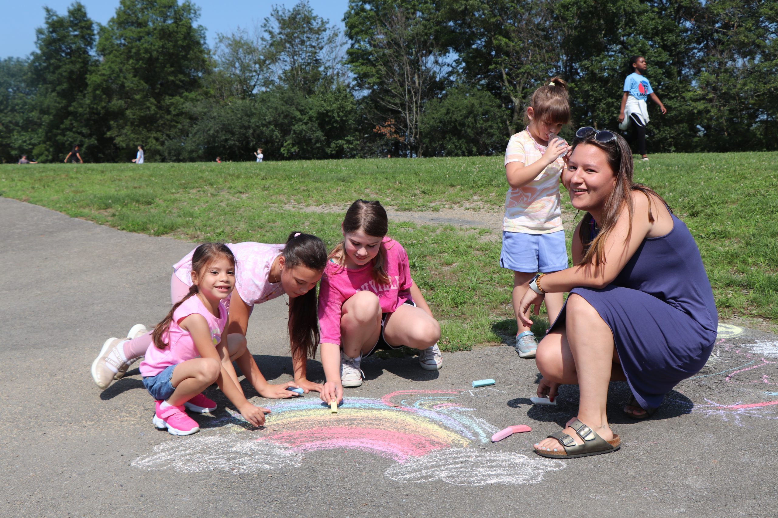 Children and an adult female draw a rainbow in colorful chalk on black asphalt.