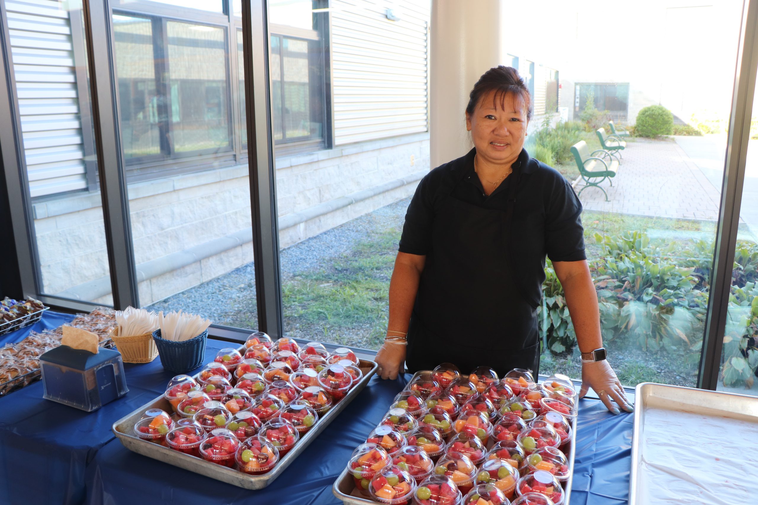 A woman wearing all black stands in front of serving trays filled with fruit in a brightly lit hallway.