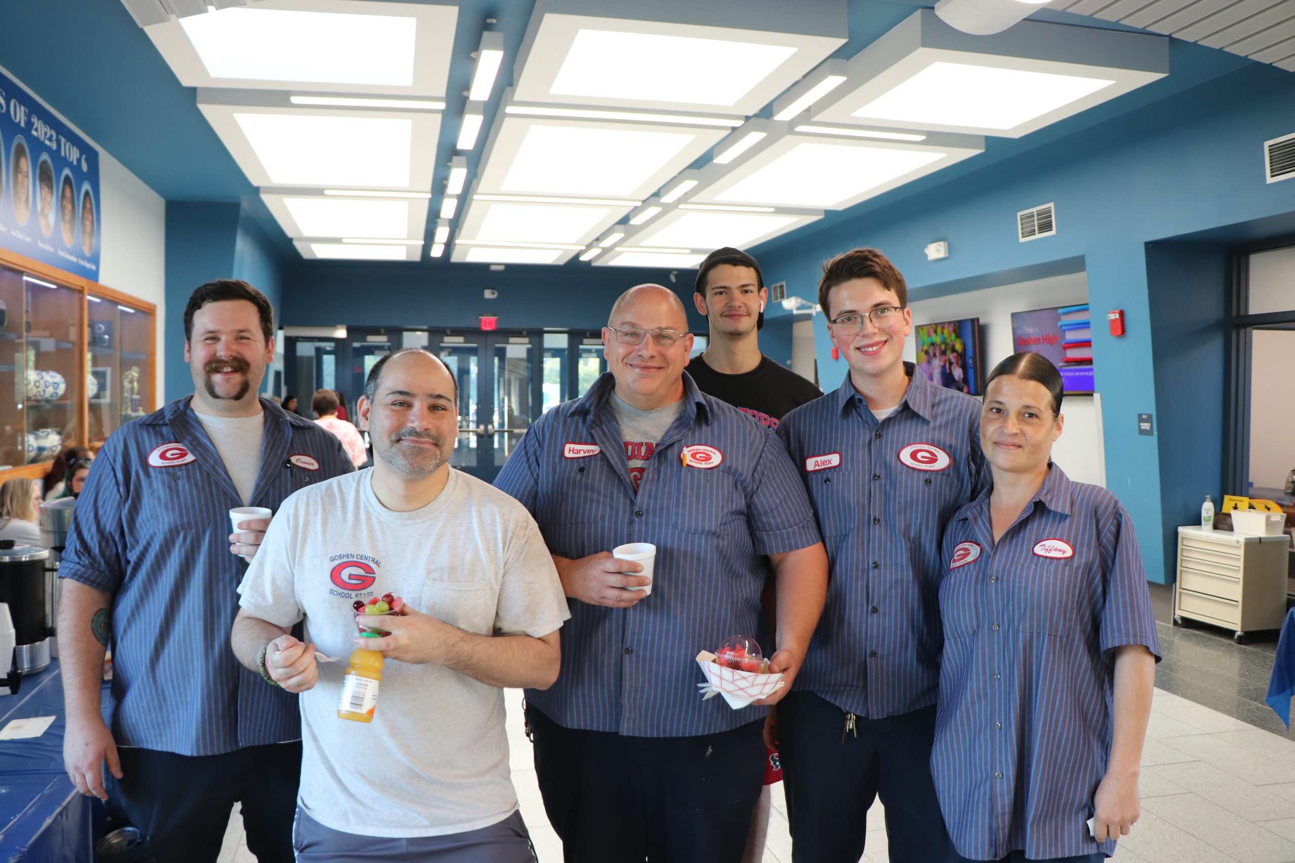 Custodians in uniform stand together smiling with fruit cups and juice.