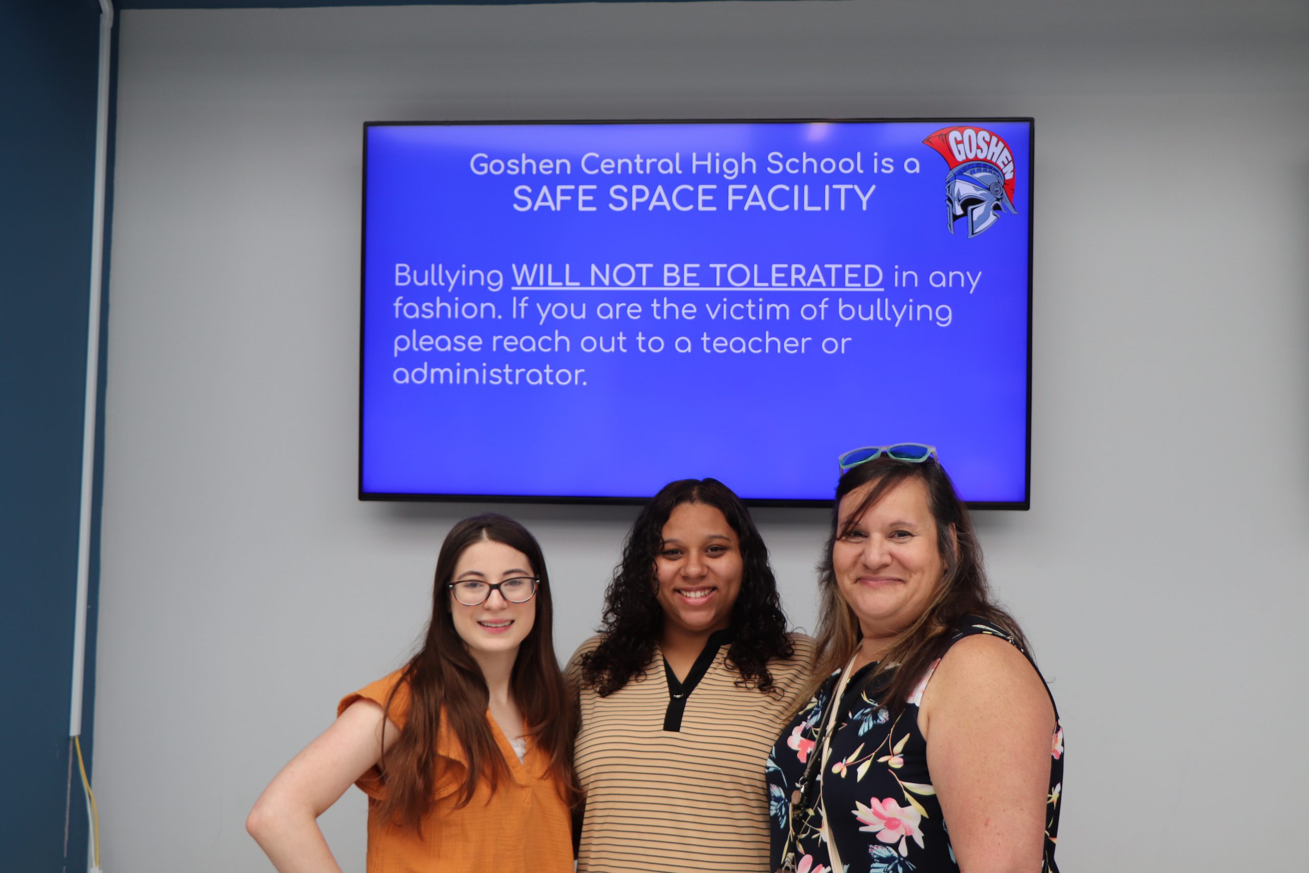 Three women stand together smiling in front of a screen that says "Goshen Central High School is a Safe Space Facility"