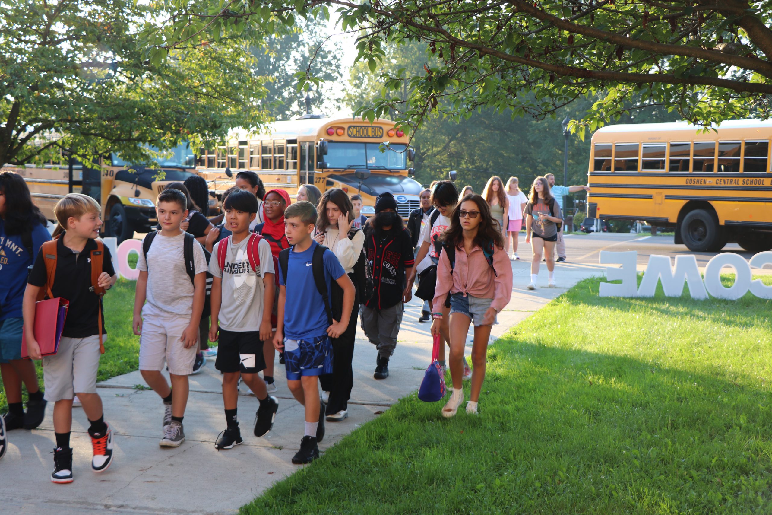 Preteens walk down a sidewalk past buses and first day of school decorations.