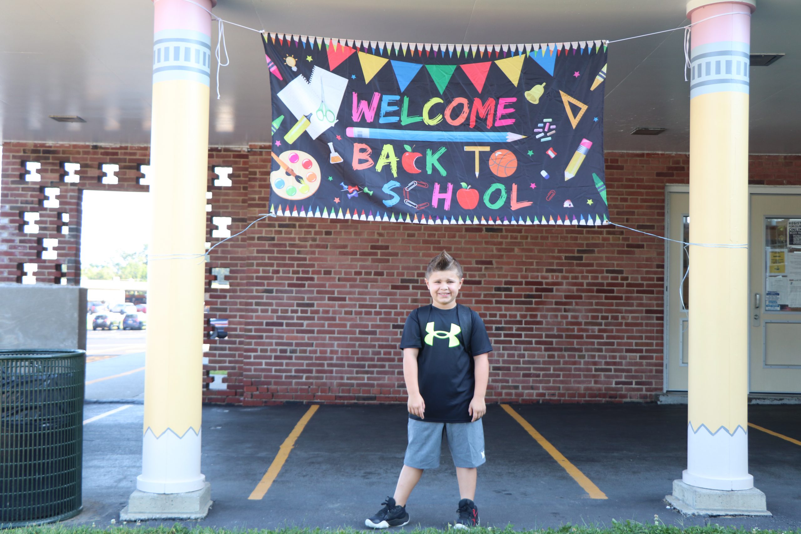 A boy smiles under a "Welcome Back to School" banner held between two pillars shaped like pencils.