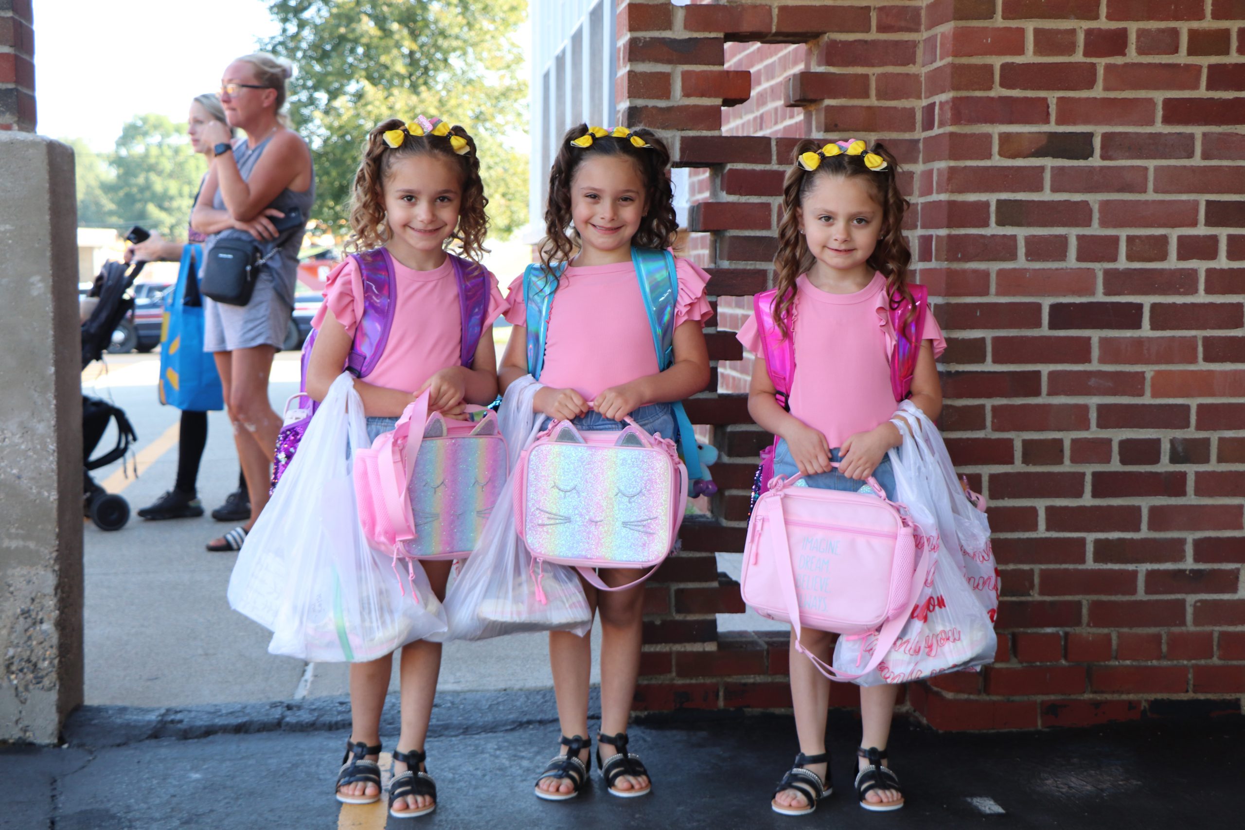 Triplets with pigtails, backpacks, lunch boxes, and plastic bags smile