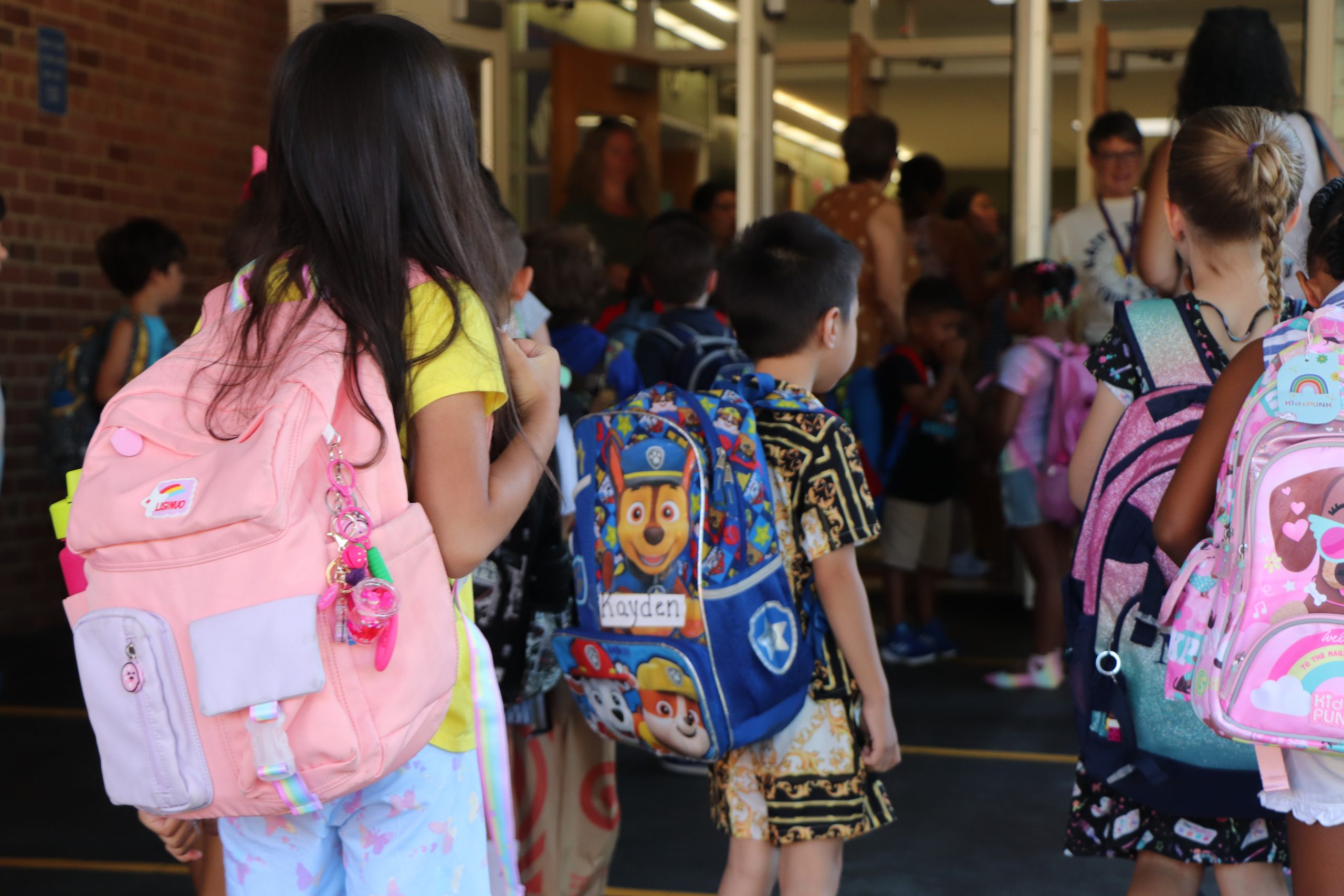 Children with backpacks walk toward a school building entrance.