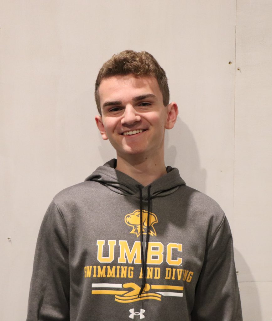 A teenager in a UMBC Swimming and Diving hoodie smiles in front of a white backdrop.