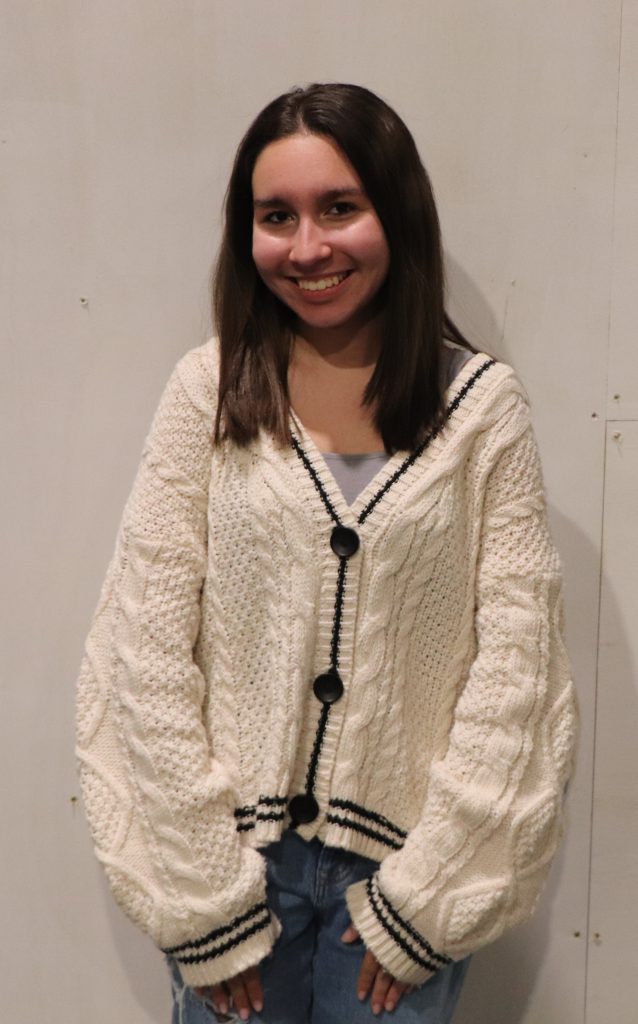 A teenager in a white knit cardigan smiles in front of a white backdrop.