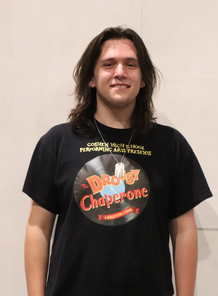 A teenager with a Goshen High School Performing Arts T-shit smiles in front of a white backdrop.