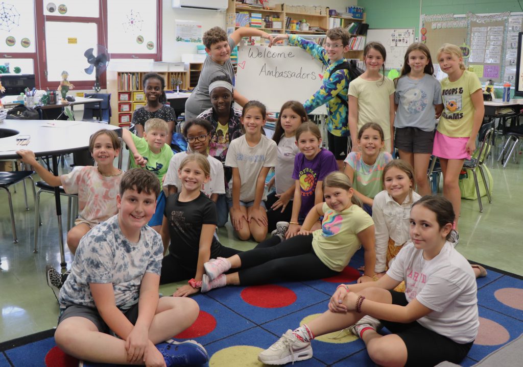Fifth graders smile and pose next to a white board that says "Welcome Ambassadors!" Two students frame the sign with arms in the shape of a heart.