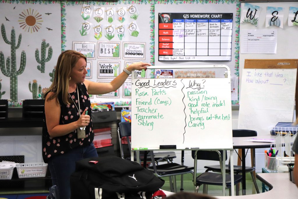 A woman stands next to a white board with two categories written, "Good Leaders" and "Why?" She is in a colorful classroom with lots of signs and materials around her.