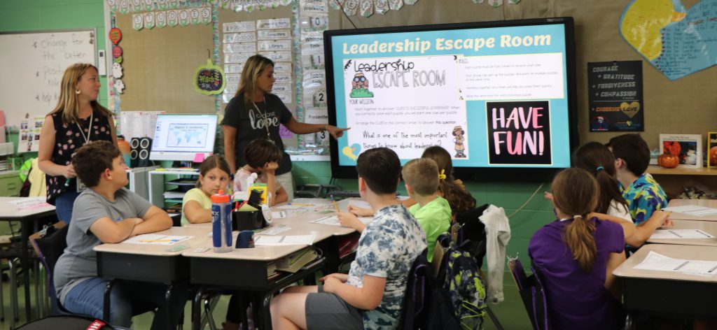 Two female teachers stand next to a presentation on a SmartBoard that reads "Leadership Escape Room" while fifth grade students sit at desks with paper and pencils.