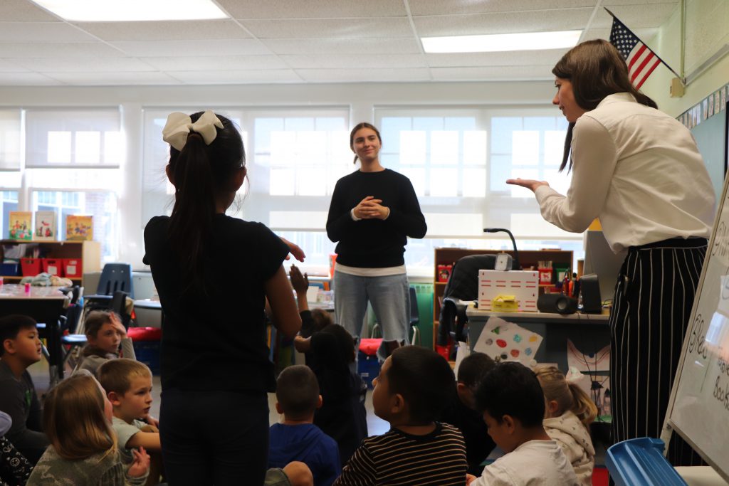 A teenage girl, female teacher, and young girl with a bow in her hair stand in a classroom surrounded by elementary students sitting together on the floor.