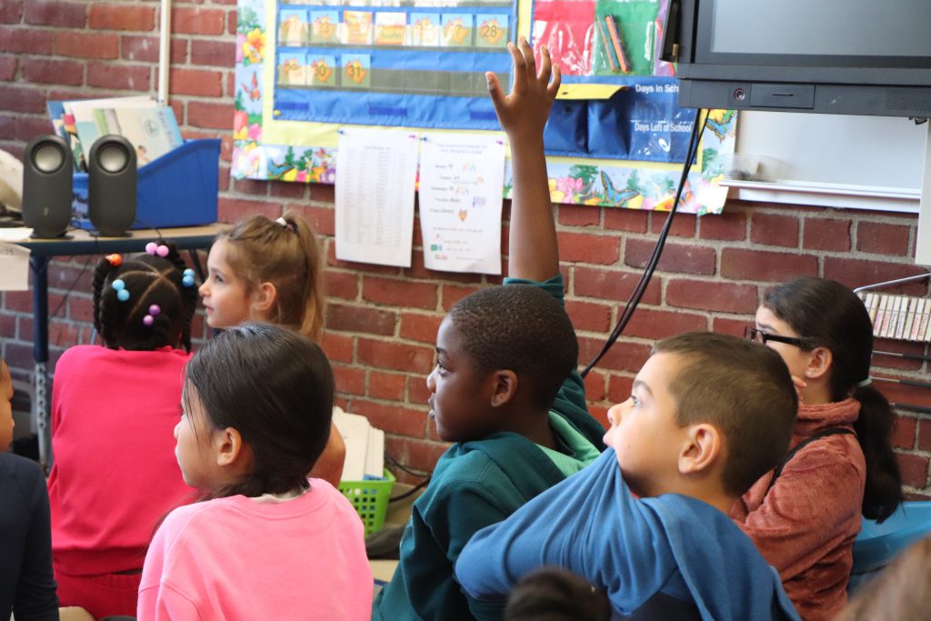 Second grade students sit on the floor of a colorful classroom facing the back of the room. One student is raising his hand.