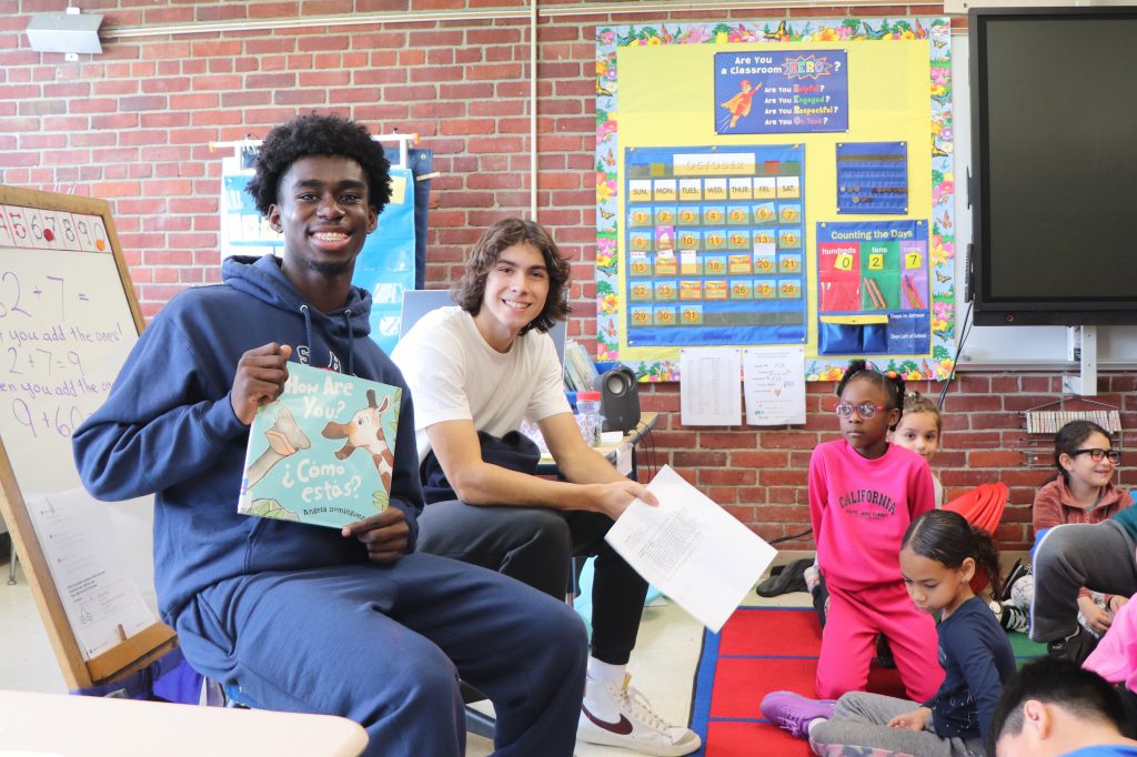Two teenagers sit with a picture book called "How are you? Como Estas?" in front of a class of second grade students sitting on the floor.