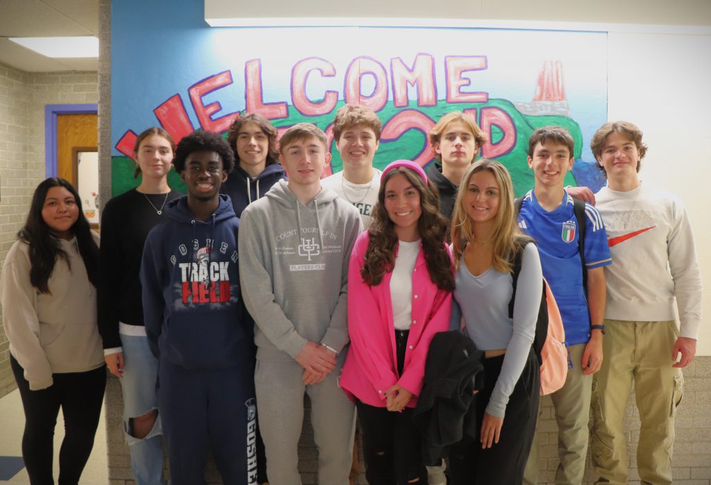 A group of teenagers stand smiling in front of a hallway mural that says "Welcome to 2nd Grade."