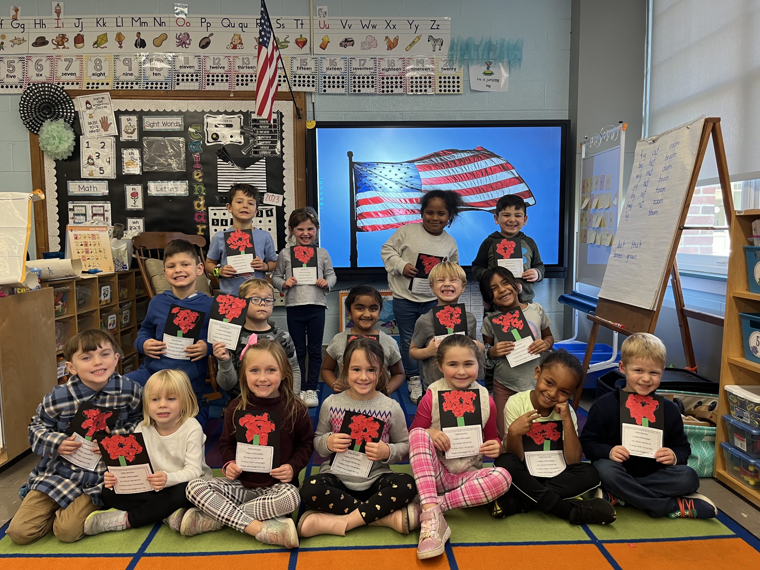 Elementary school students sit together on a carpet in their classroom in front of an American flag with handmade Veterans Day cards.