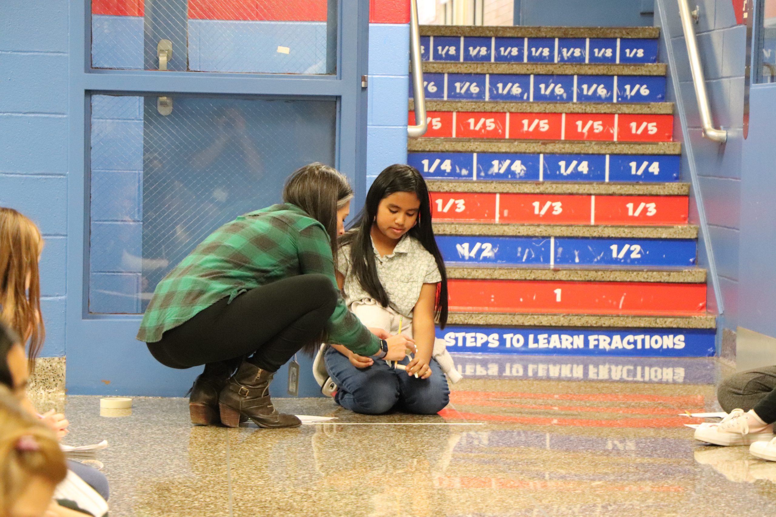 An elementary school student and a teacher work together to launch a small handmade turkey from a spoon down a hallway.