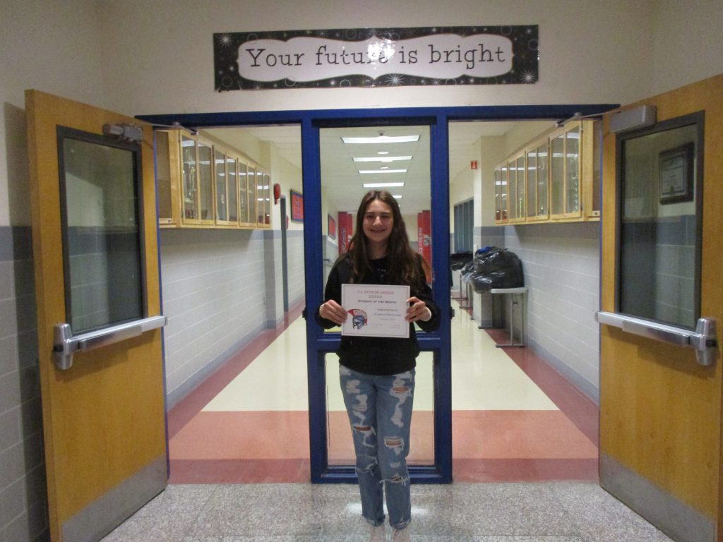 A middle school student smiles holding a certificate in front of hallway doors with the text "Your future is bright" above her.