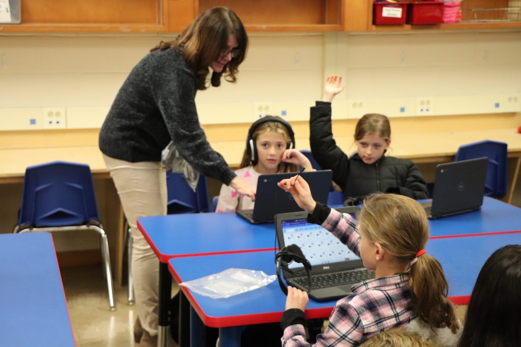 A teacher points something out on a Chromebook to an elementary school student wearing headphones at a blue table. A student next to her raises her hand.