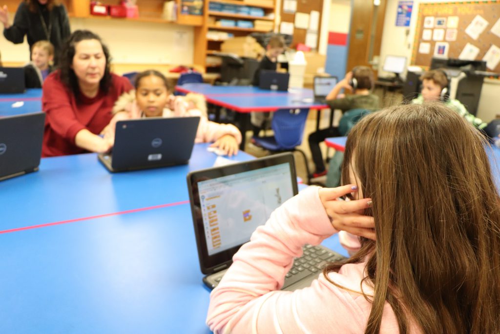 A teacher works with an elementary school student on a Chromebook across from another student at a blue table during the last session of the Coding Club at Goshen Intermediate School.