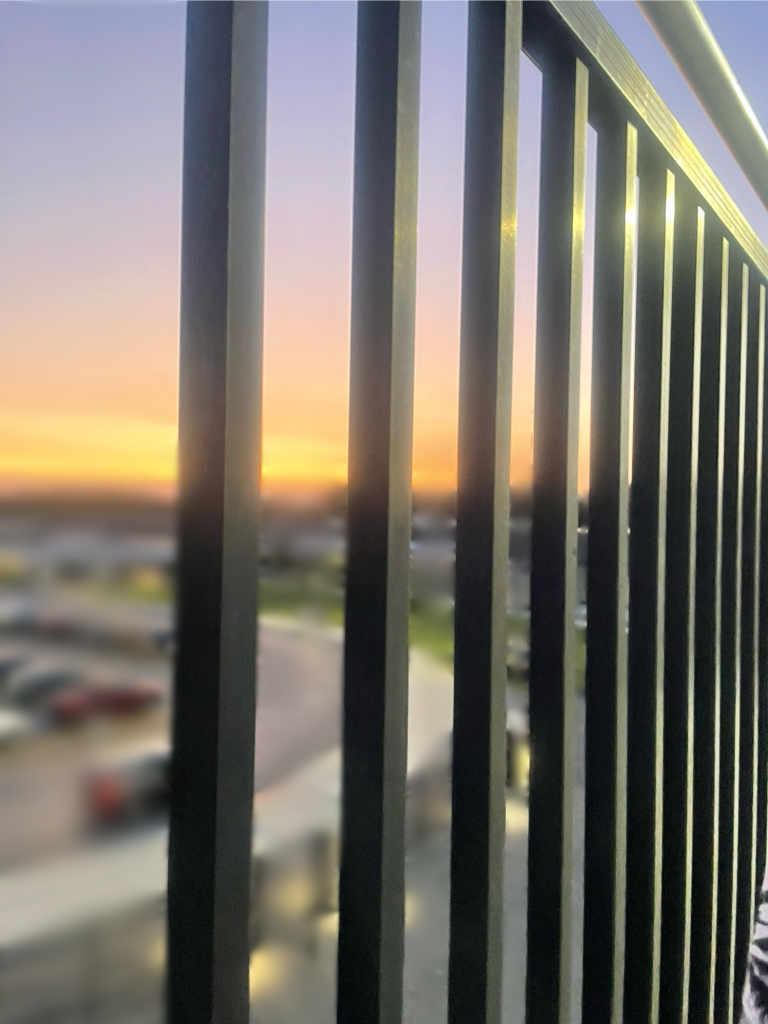 A photograph of a pink, orange and purple sunset is out of focus behind iron bars of some sort of balcony.