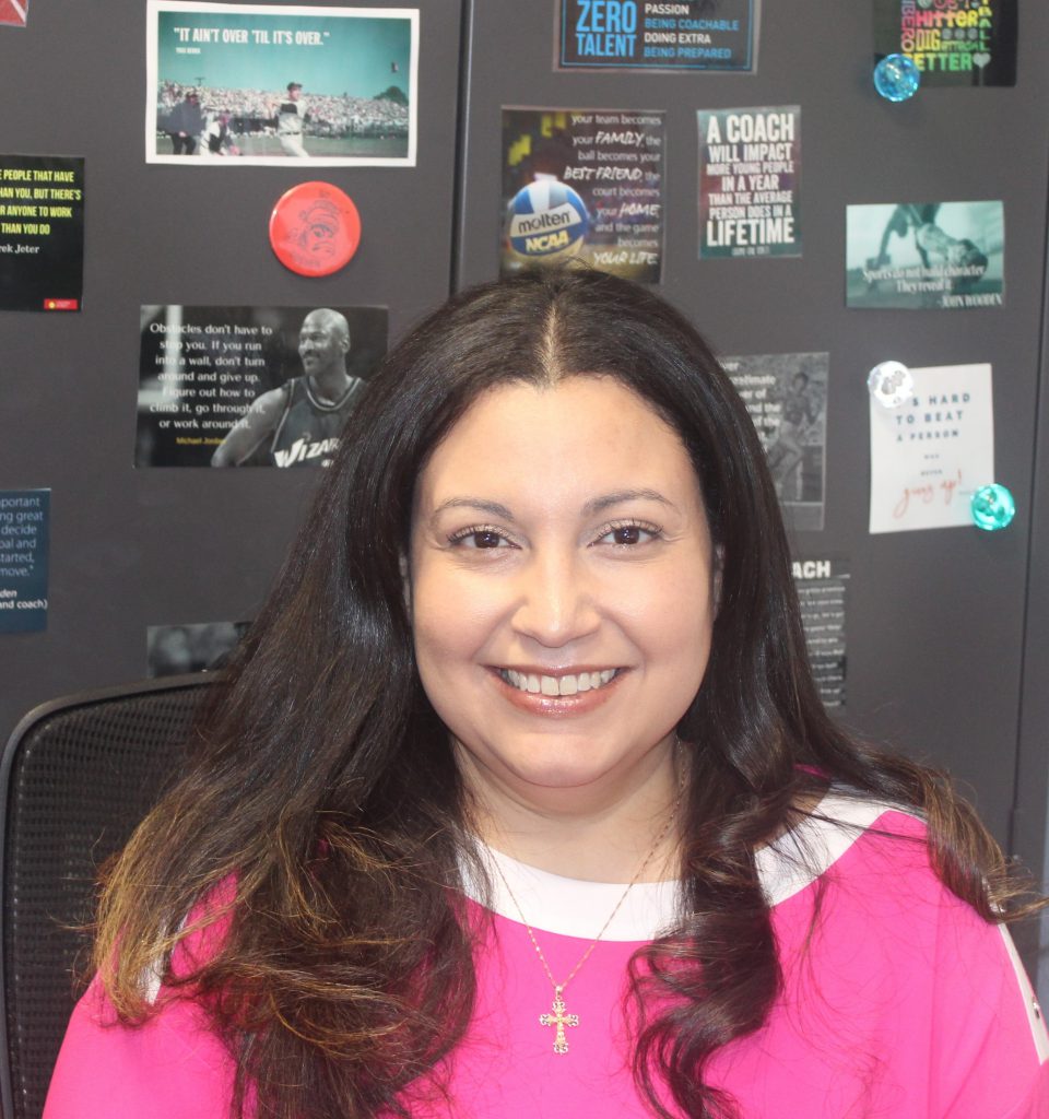 A photo of a woman in a pink shirt with a cross necklace in front of lockers with inspirational athletic quotations.