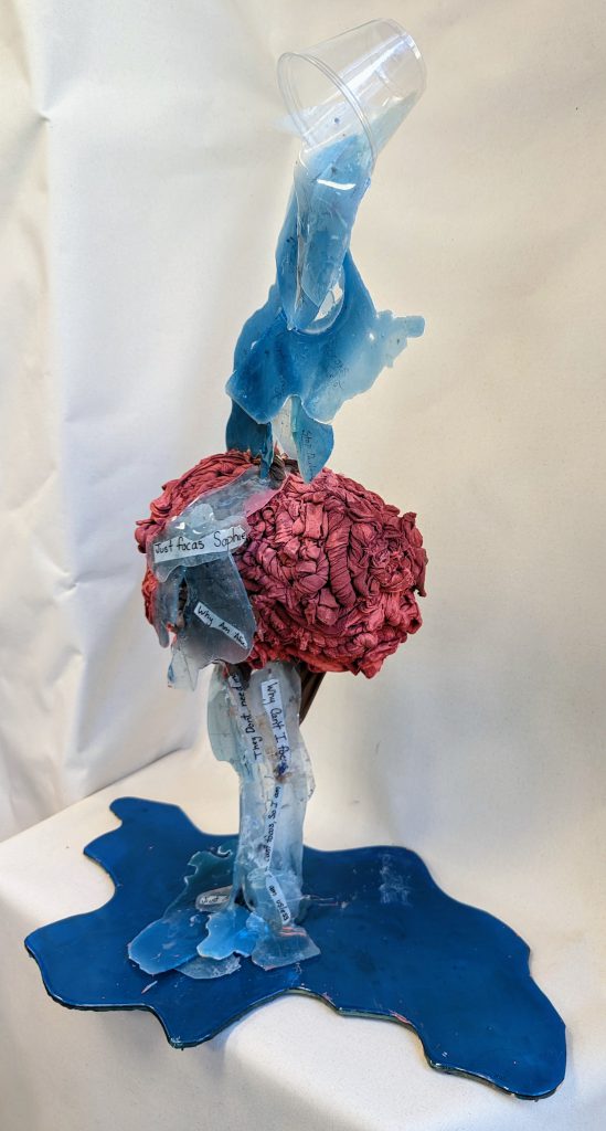 Sculpture of what looks like a pink/red brain with blue liquid being poured out of a glass onto it and spilling below.