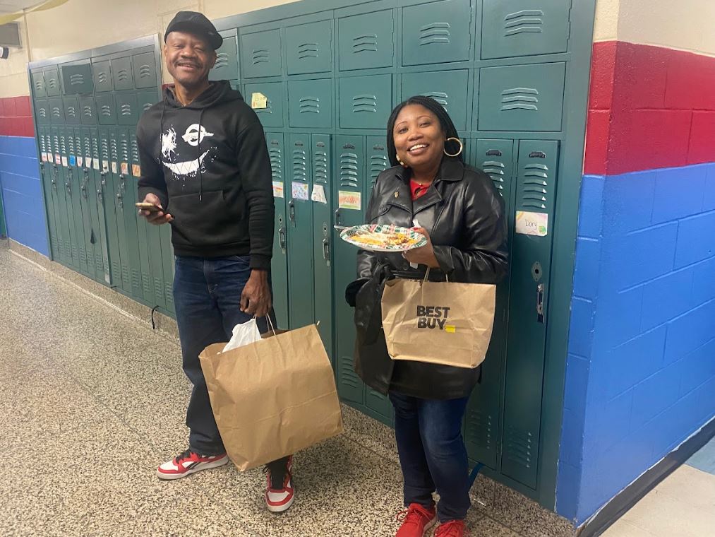 Two adults with paper plates of food from different cultures smile for the camera in the hallway at GIS during the 5th Grade Culture Fest.