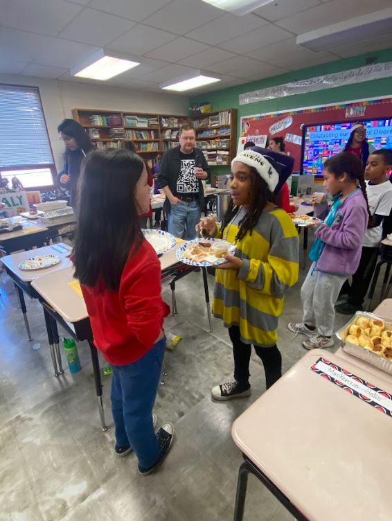 Elementary school students and adults with paper plates of food mingle in a classroom in front of informative trifolds depicting various cultures in the Western Hemisphere.