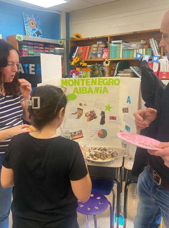 Elementary school students and adults with paper plates of food mingle in a classroom in front of informative trifolds depicting the cultures of Guatemala and Montenegro/Albania.