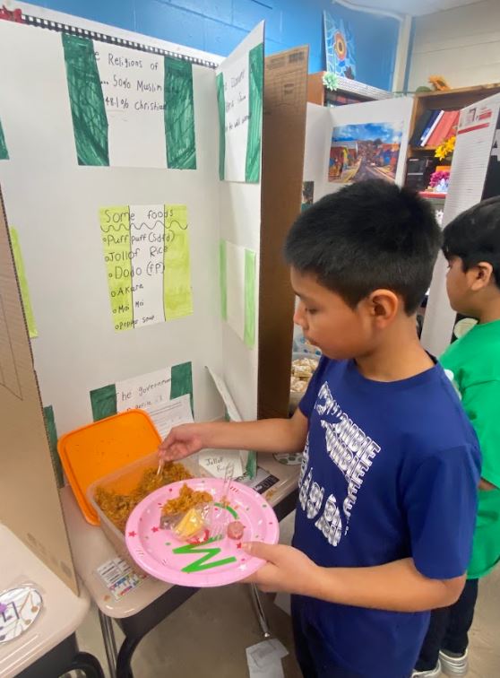 Elementary school students with paper plates of food mingle in a classroom in front of informative trifolds depicting various cultures in the Western Hemisphere.
