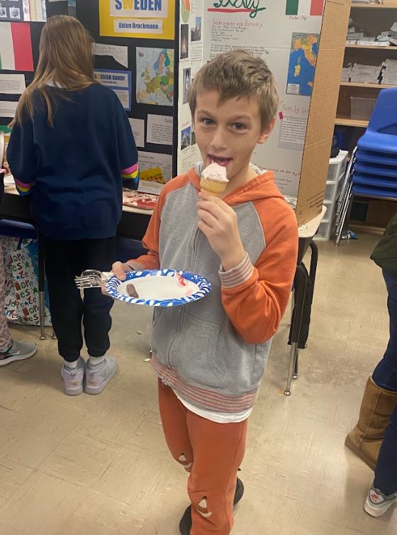 An elementary school student holds a paper plate in one hand and an ice cream cone in the other, standing in front of student-made trifolds with information on Sicily and Sweden.