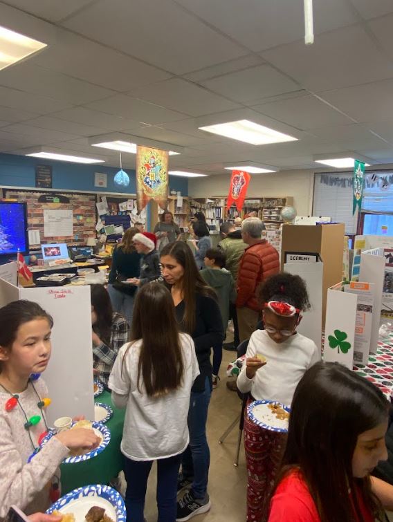 Elementary school students and adults with paper plates of food mingle in a classroom in front of informative trifolds depicting various cultures in the Western Hemisphere.