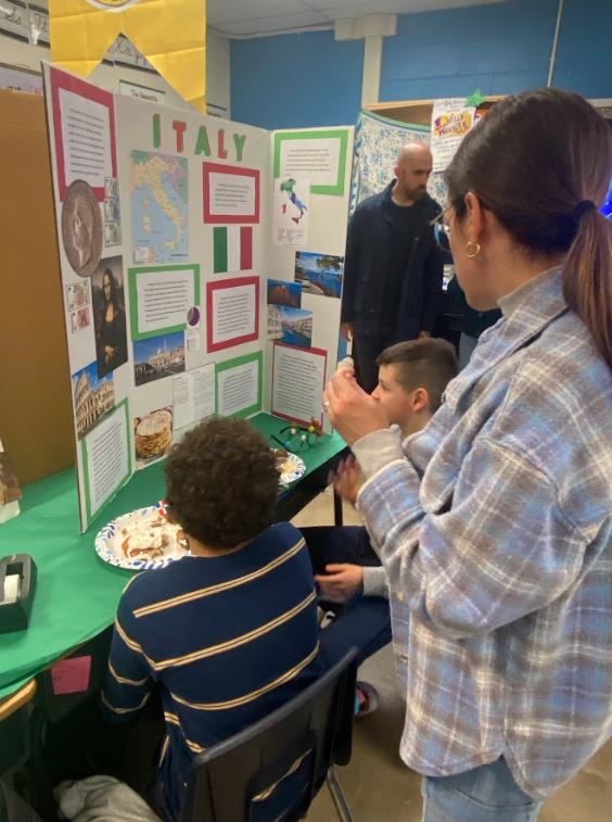 Elementary school students and adults with paper plates of food mingle in a classroom in front of an informative trifold depicting Italian culture.