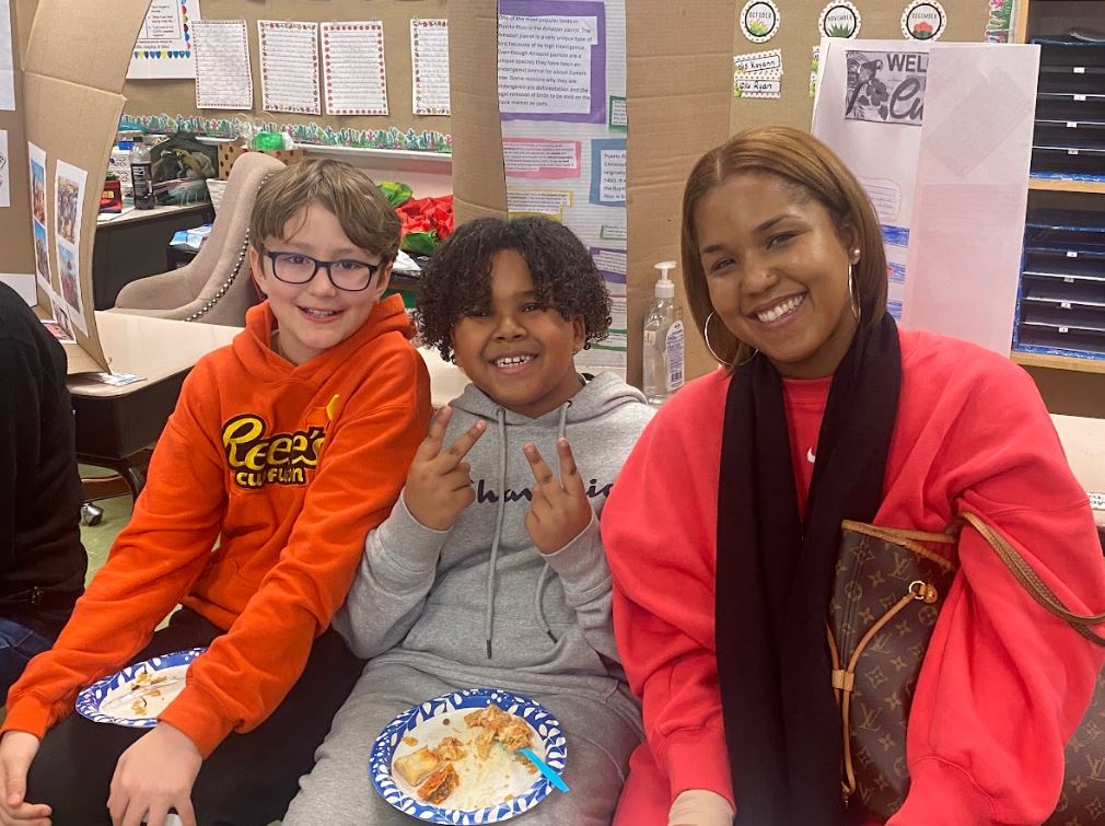 Elementary school students with paper plates of food and an adult smile for the camera in front of informative trifolds depicting various cultures in the Western Hemisphere.