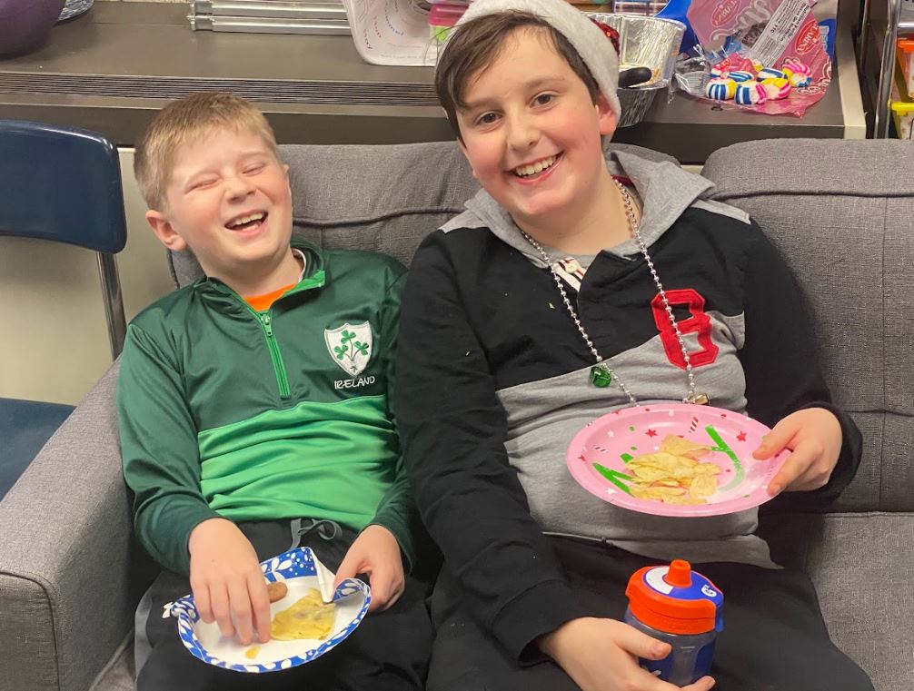 Two elementary school students in sweaters smile on a couch with paper plates of food.