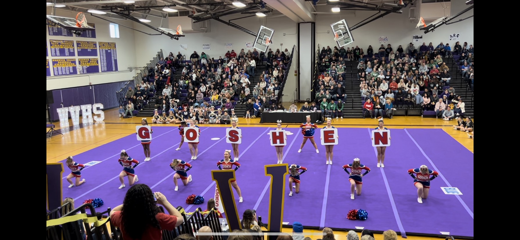 Cheerleaders hold up letters spelling out "Goshen" in a gym with a purple floor and crowded bleachers. 