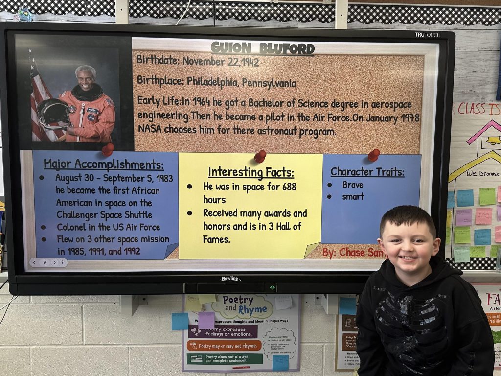 An elementary school student stands next to a slide with research on Guion Bluford.