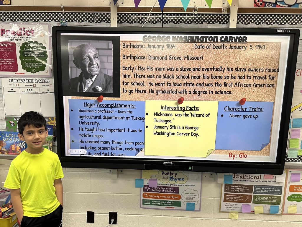 An elementary school student stands next to a slide with research on George Washington Carver.