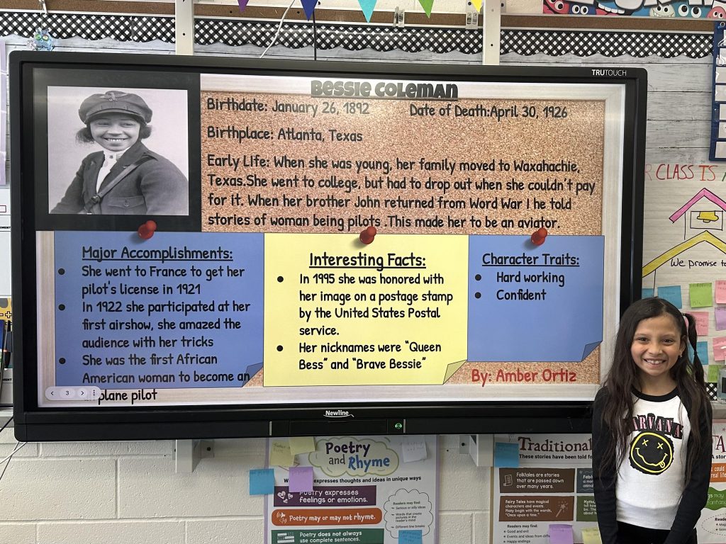 An elementary school student stands next to a slide with research on Bessie Coleman.