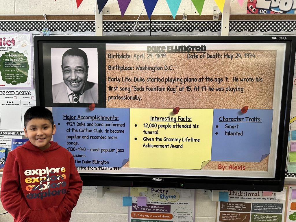 An elementary school student stands next to a slide with research on Duke Ellington.
