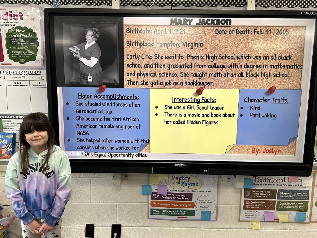 An elementary school student stands next to a slide with research on Mary Jackson.