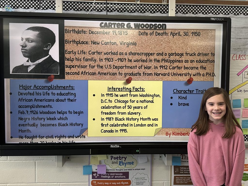An elementary school student stands next to a slide with research on Carter G. Woodson.