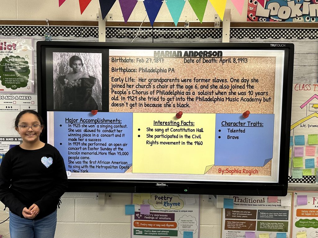 An elementary school student stands next to a slide with research on Marian Anderson.