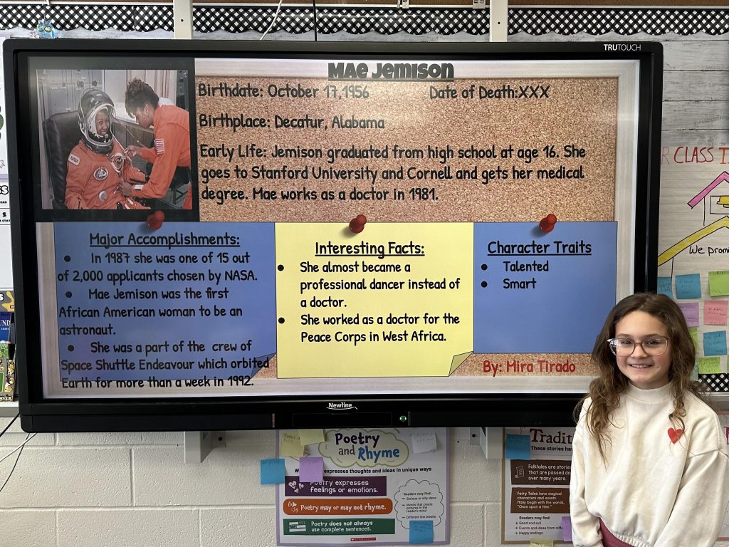 An elementary school student stands next to a slide with research on Mae Jemison.