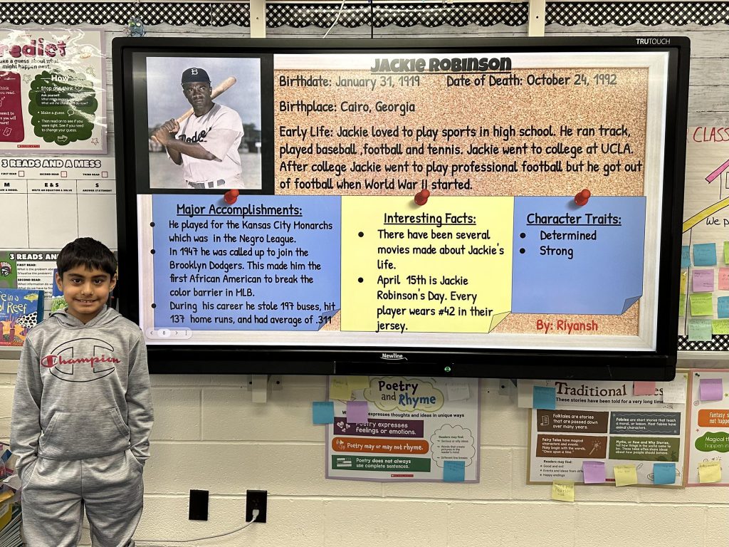 An elementary school student stands next to a slide with research on Jackie Robinson.