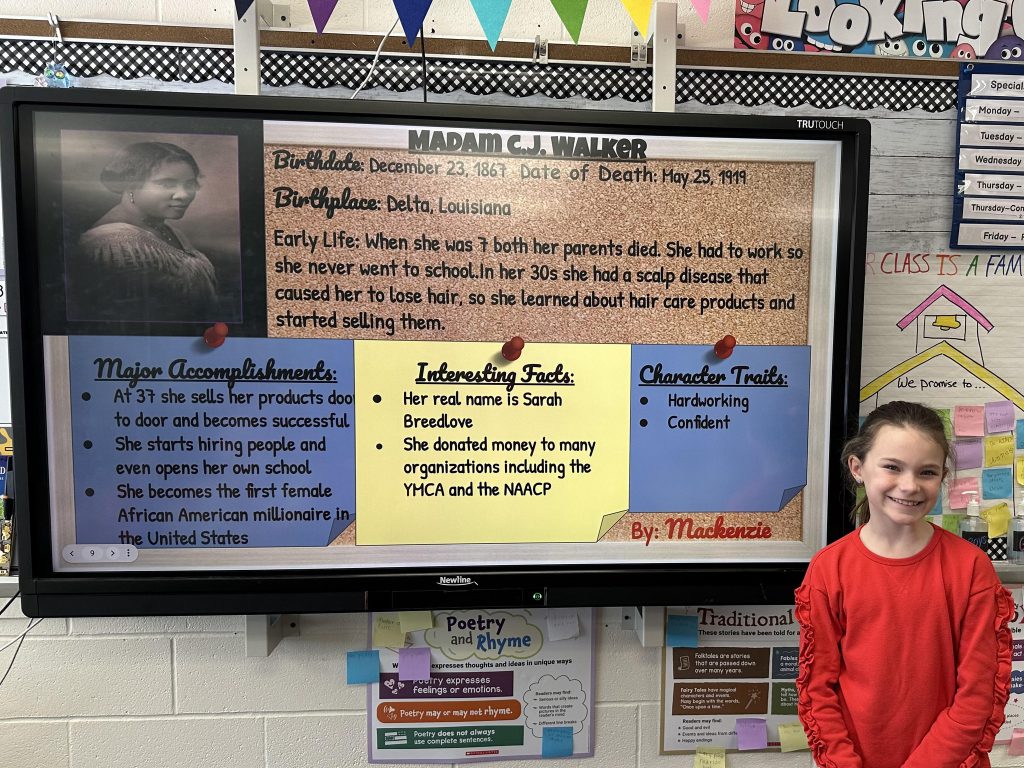 An elementary school student stands next to a slide with research on Madam C.J. Walker.