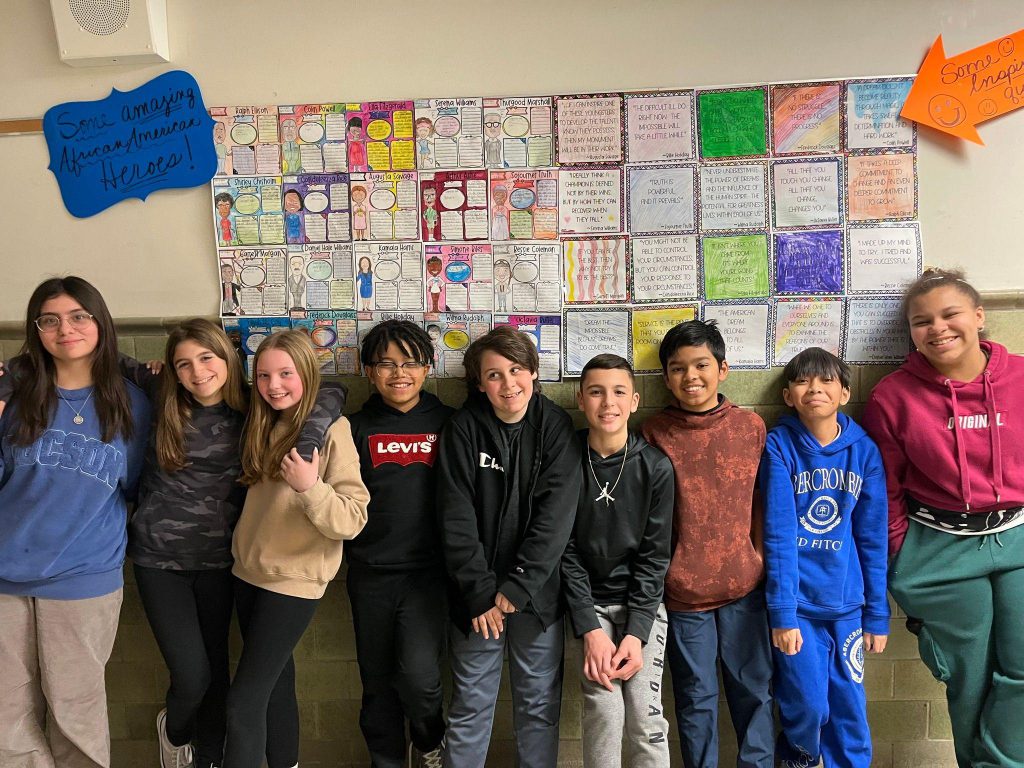 Middle school students smile together in front of a handmade paper quilt with images of famous Black Americans and their quotes with the text "Some amazing African American Heroes!" next to it. 
