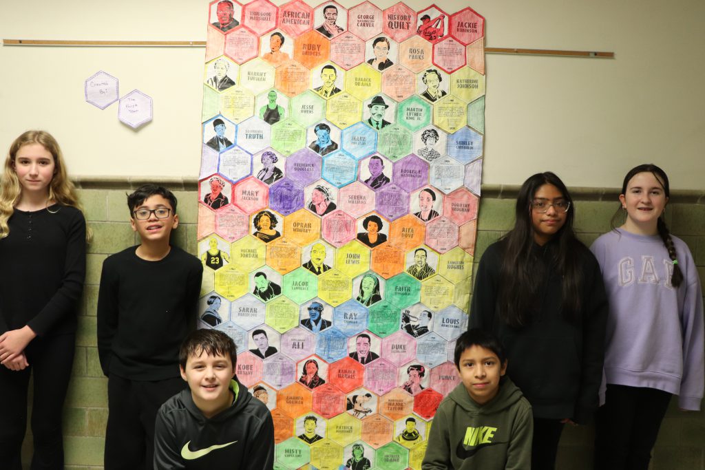 Middle school students stand next to a rainbow-colored, handmade tessellated paper quilt with images of famous Black Americans and their accomplishments.