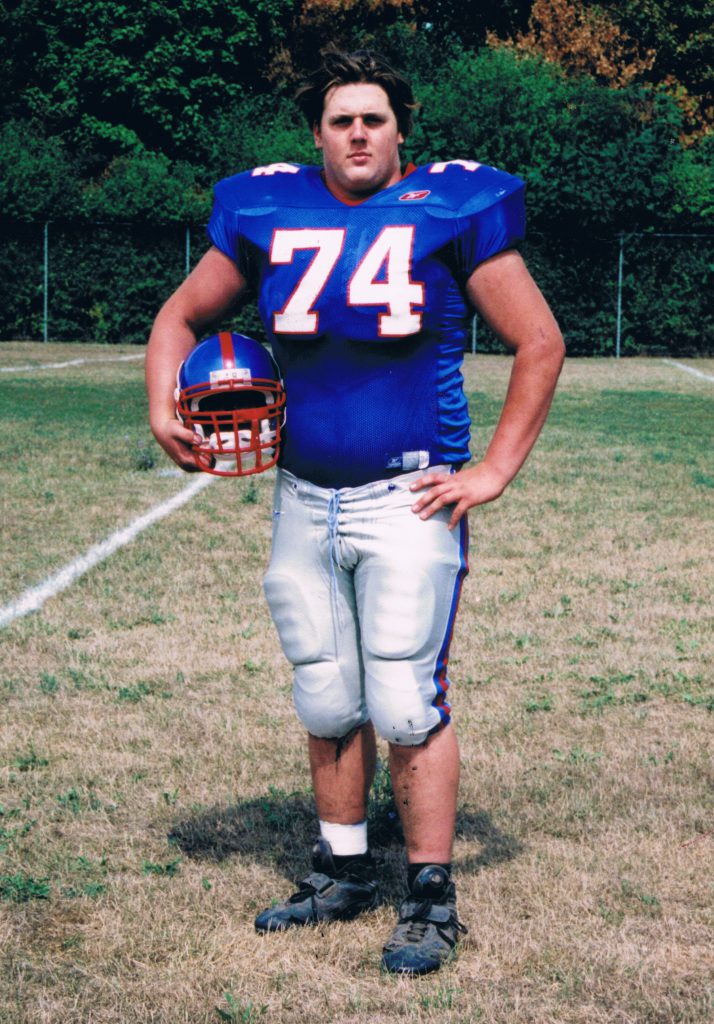A young man in a Goshen Football team uniform with the number 74 on the front poses with his helmet.