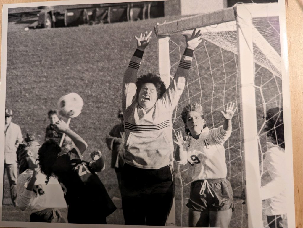 A girl in soccer gear jumps up to block a goal in a black-and-white photo with teammates and a big goal net beside her.
